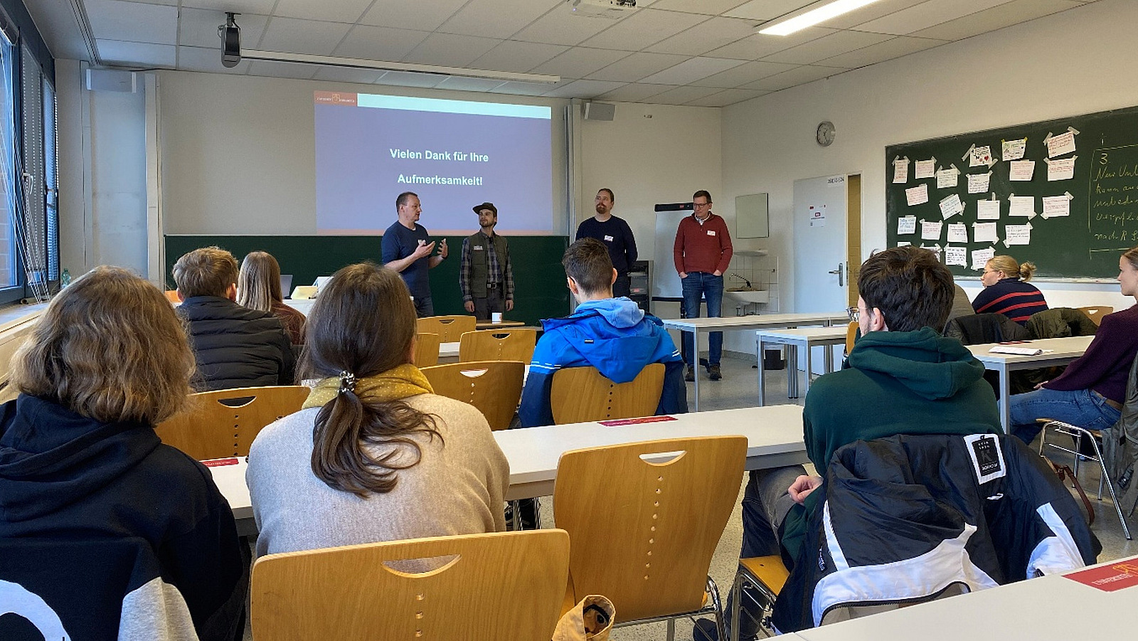 Four men are standing in a seminar room where pupils are sitting.