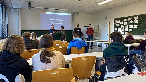 Four men are standing in a seminar room where pupils are sitting.