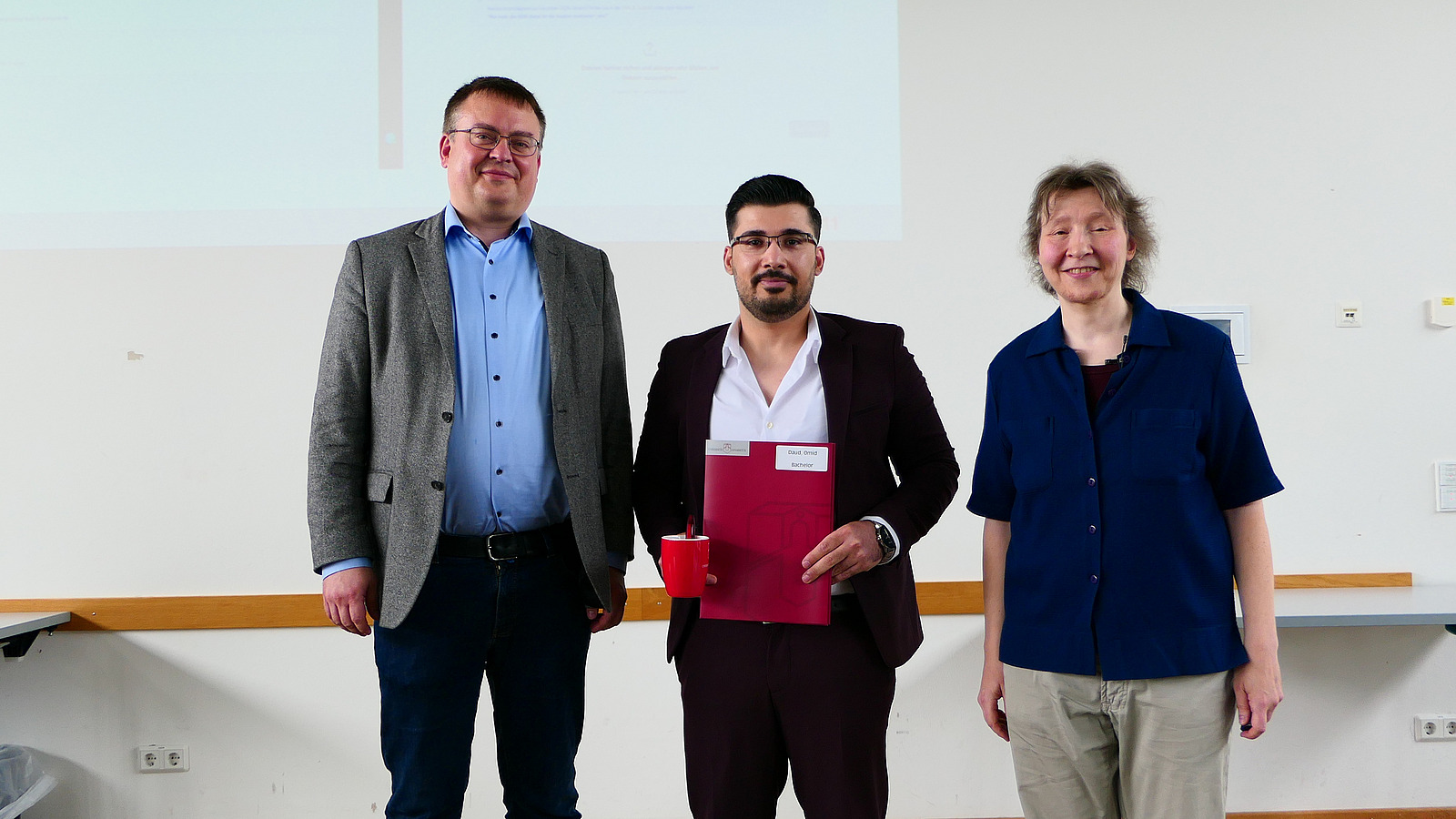 Ein Student mit Studiendekanin und Dekan stehen nebeneinander. Der Student hat eine Mappe in der Hand