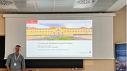 Gerrit Wittke, im Hintergrund erste Folie seines Vortrags, auf der das Universitätsgebäude Osnabrück, gelbes Schloss, sowie das Thema seines Vortrags „Conditional Tail-Risk in Social Trading“ abgebildet sind.  