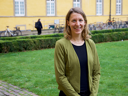 Eine Person mit halblangen braunen Haaren in einem schwarzen Kleid und grüner Jacke steht vor einem historischen Gebäude uns lächelt in die Kamera.