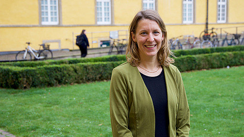Eine Person mit halblangen braunen Haaren in einem schwarzen Kleid und grüner Jacke steht vor einem historischen Gebäude uns lächelt in die Kamera.