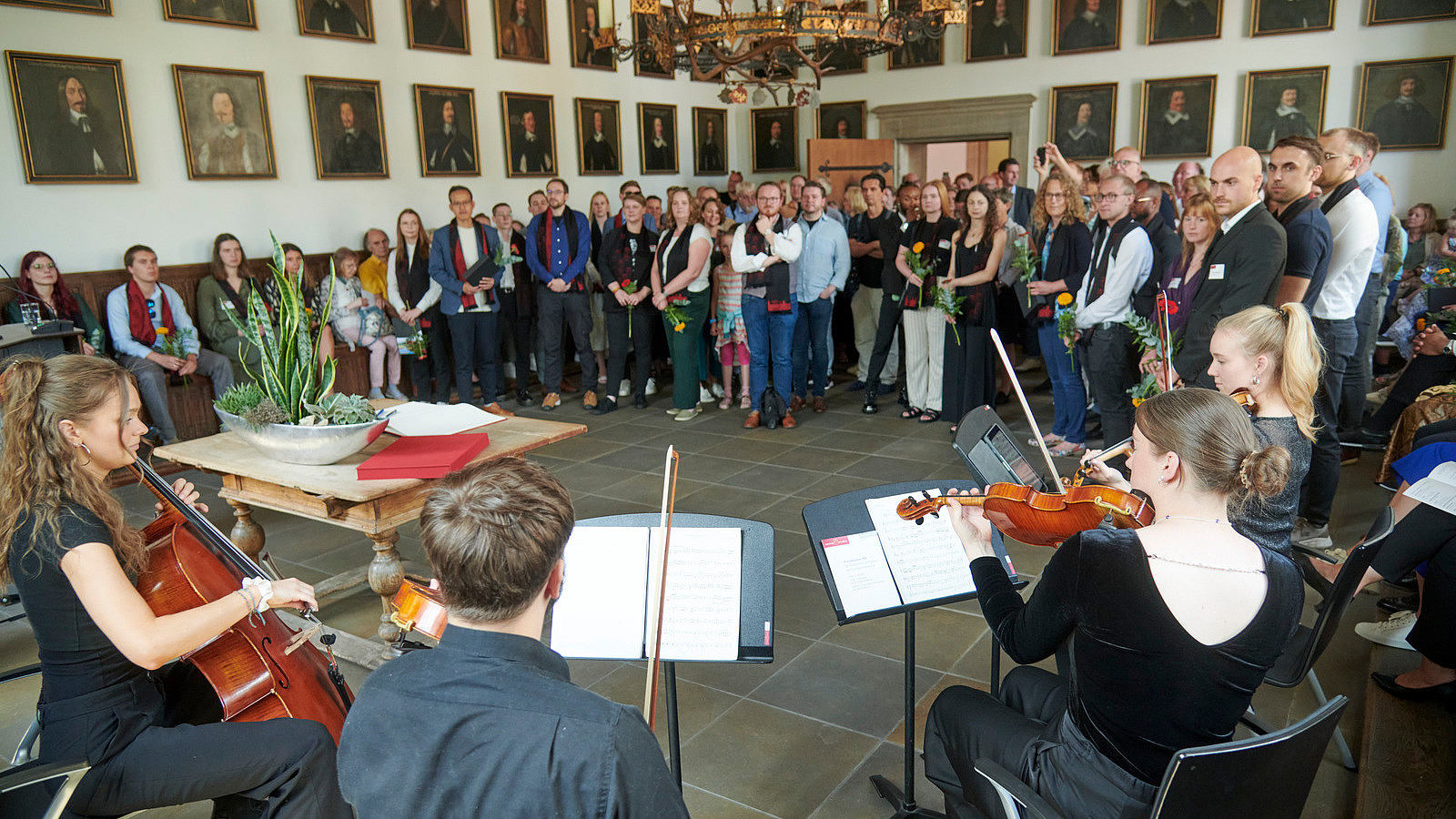Numerous people stand in a semi-circle in a room with a historical feel. Four people with string instruments are sitting in front of them