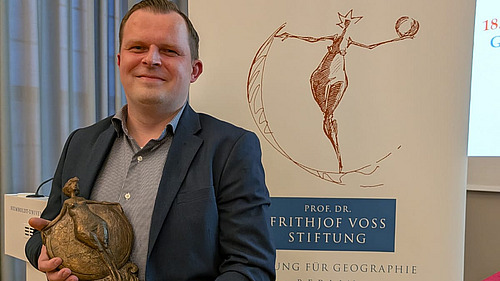 Ein Mann steht vor einem Plakat und hält eine kleine Statue in der Hand. 