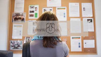 Eine Person steht mit dem Rücken zur Kamera und blickt auf ein Informationsbrett. Auf dem Brett sind mehrere Plakate und Mitteilungen angebracht, die Informationen und Fotos enthalten.