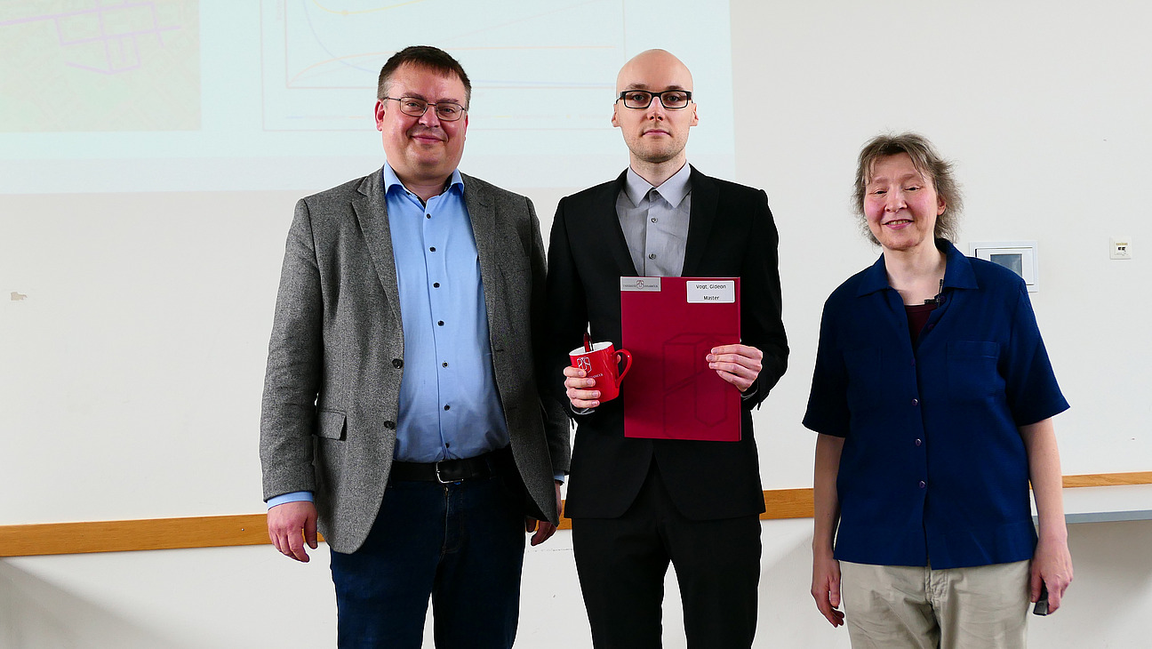 Ein Student mit Studiendekanin und Dekan stehen nebeneinander. Der Student hat eine Mappe in der Hand