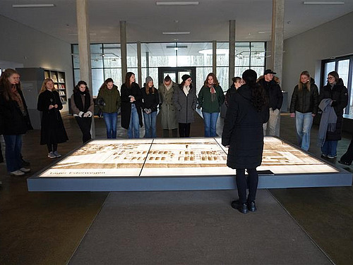 Eine Gruppe von Menschen betrachtet eine große Darstellung auf dem Boden in einem hellen Ausstellungsraum. Die Personen, hauptsächlich junge Frauen, scheinen die Details des Objekts zu diskutieren.