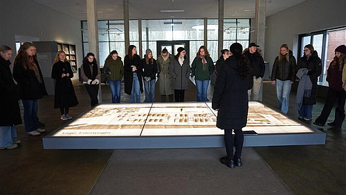 Eine Gruppe von Menschen betrachtet eine große Darstellung auf dem Boden in einem hellen Ausstellungsraum. Die Personen, hauptsächlich junge Frauen, scheinen die Details des Objekts zu diskutieren.