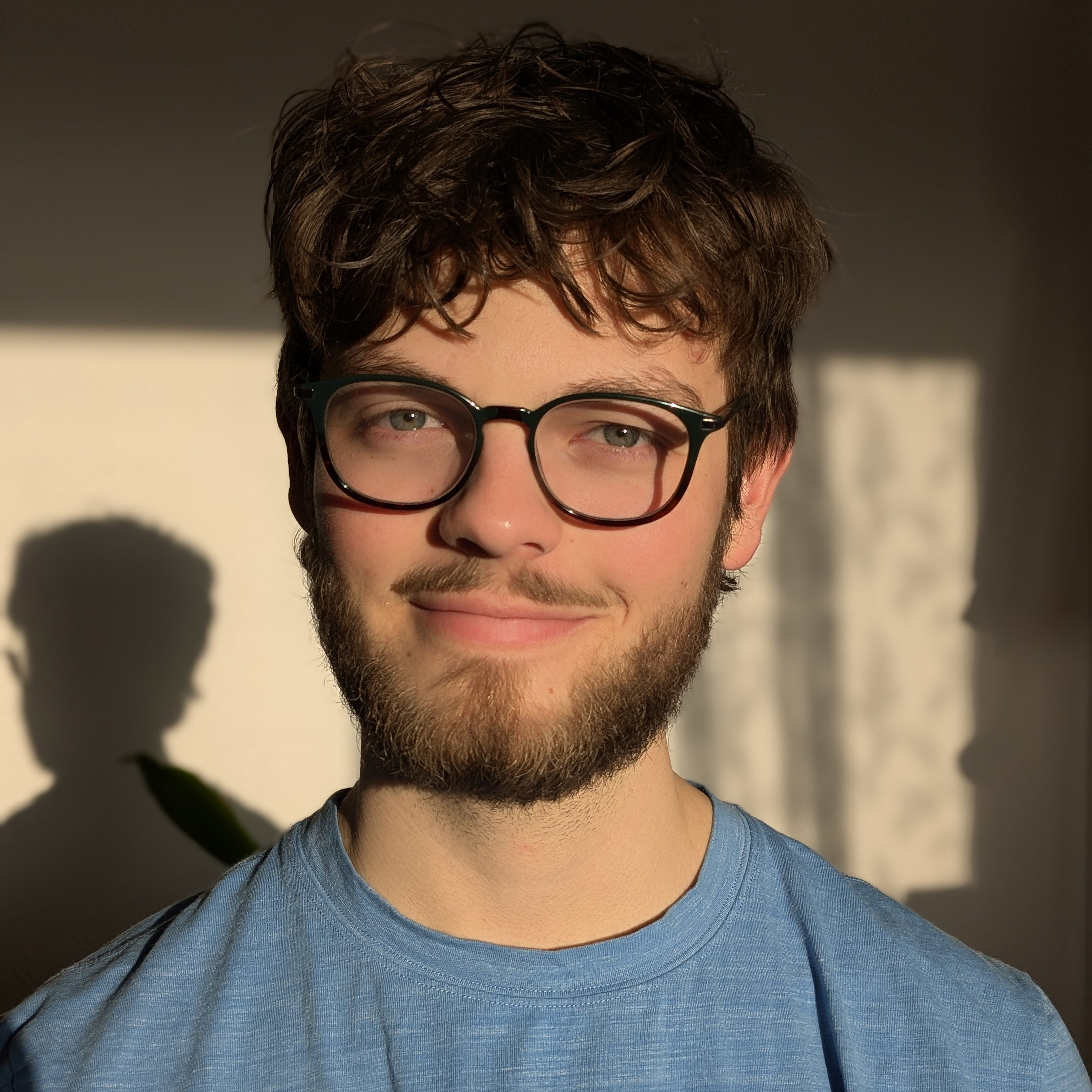 Portrait von einem jungen Mann mit braunen Haaren. Er tr&auml;gt eine schwarze Brille und ein hellblaues T-Shirt.