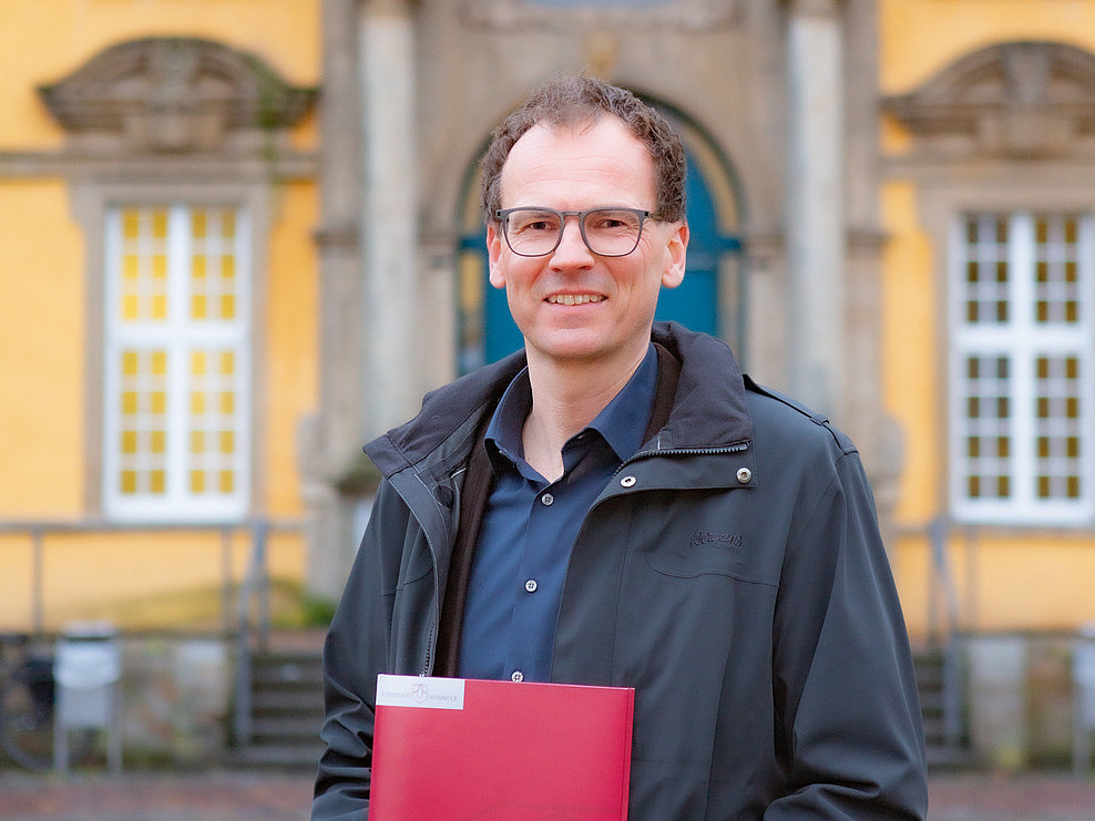 Prof. Woppowa steht im Innenhof des Osnabrücker Schlosses. Er lächelt in die Kamera. In der Hand hält er eine rote Mappe mit dem Logo der Universität Osnabrück