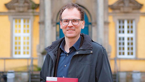 Prof. Woppowa steht im Innenhof des Osnabr&uuml;cker Schlosses. Er l&auml;chelt in die Kamera. In der Hand h&auml;lt er eine rote Mappe mit dem Logo der Universit&auml;t Osnabr&uuml;ck
