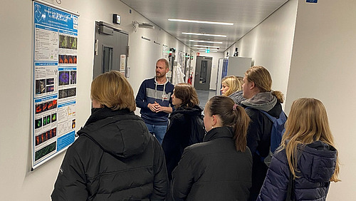 A man and a group of pupils are standing in a corridor in front of a poster.