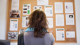 Eine Person steht mit dem Rücken zur Kamera und blickt auf ein Informationsbrett. Auf dem Brett sind mehrere Plakate und Mitteilungen angebracht, die Informationen und Fotos enthalten.