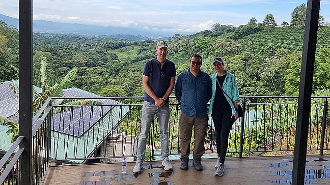 The picture shows a picturesque view from a vantage point where two people are standing on a wooden platform or balcony. The view is of a lush landscape with lots of greenery and hills in the background.