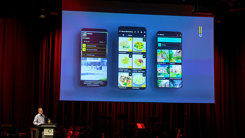 A person is standing at a lectern on stage. Above him, three smartphones displaying the OSnabrück Student Services app can be seen on the screen.