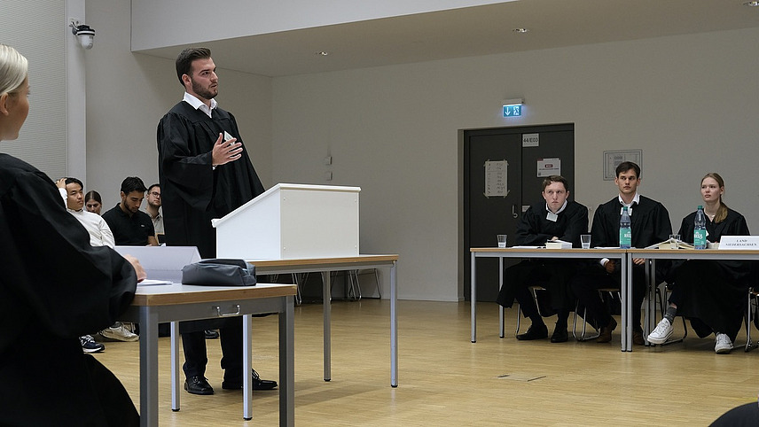 Ein Vortragender in schwarzer Robe an einem Rednerpult, mit Teams rechts und links an Tischen sitzend und Publikum im Hintergrund