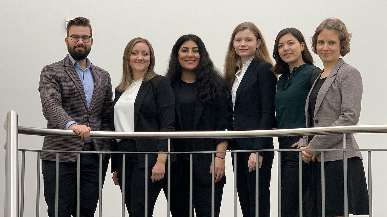 Ein Mann und f&uuml;nf Frauen in einer Reihe an halbhohen Metallgel&auml;nder stehend