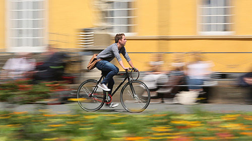 Ein junger Mann fährt auf einem schwarzen Fahrrad. Die Bewegung des Fahrrads wird durch eine Verwischung im Hintergrund deutlich, die Menschen und eine gelbe Fassade zeigt. Bunte Blumen sind im Vordergrund zu sehen.