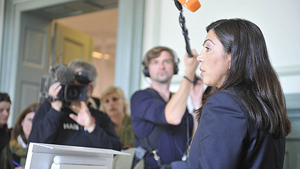 Eine Frau in formeller Kleidung spricht in ein Mikrofon während einer Pressekonferenz. Im Hintergrund sind mehrere Journalisten und Kameraleute zu sehen, wobei eine Person Videoaufnahmen macht. 