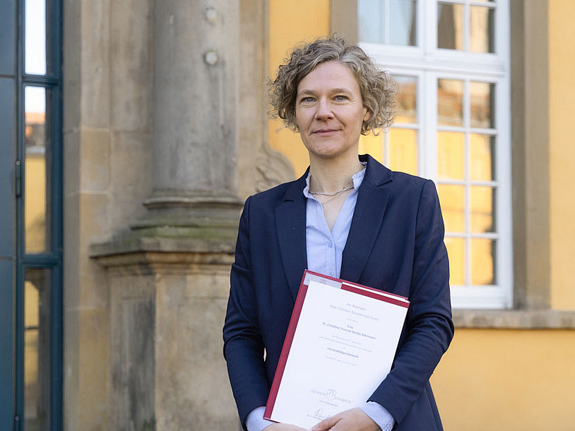 Frau im Anzug steht auf der Treppe vor dem Eingang eines gro&szlig;en Geb&auml;udes und schaut in die Kamera