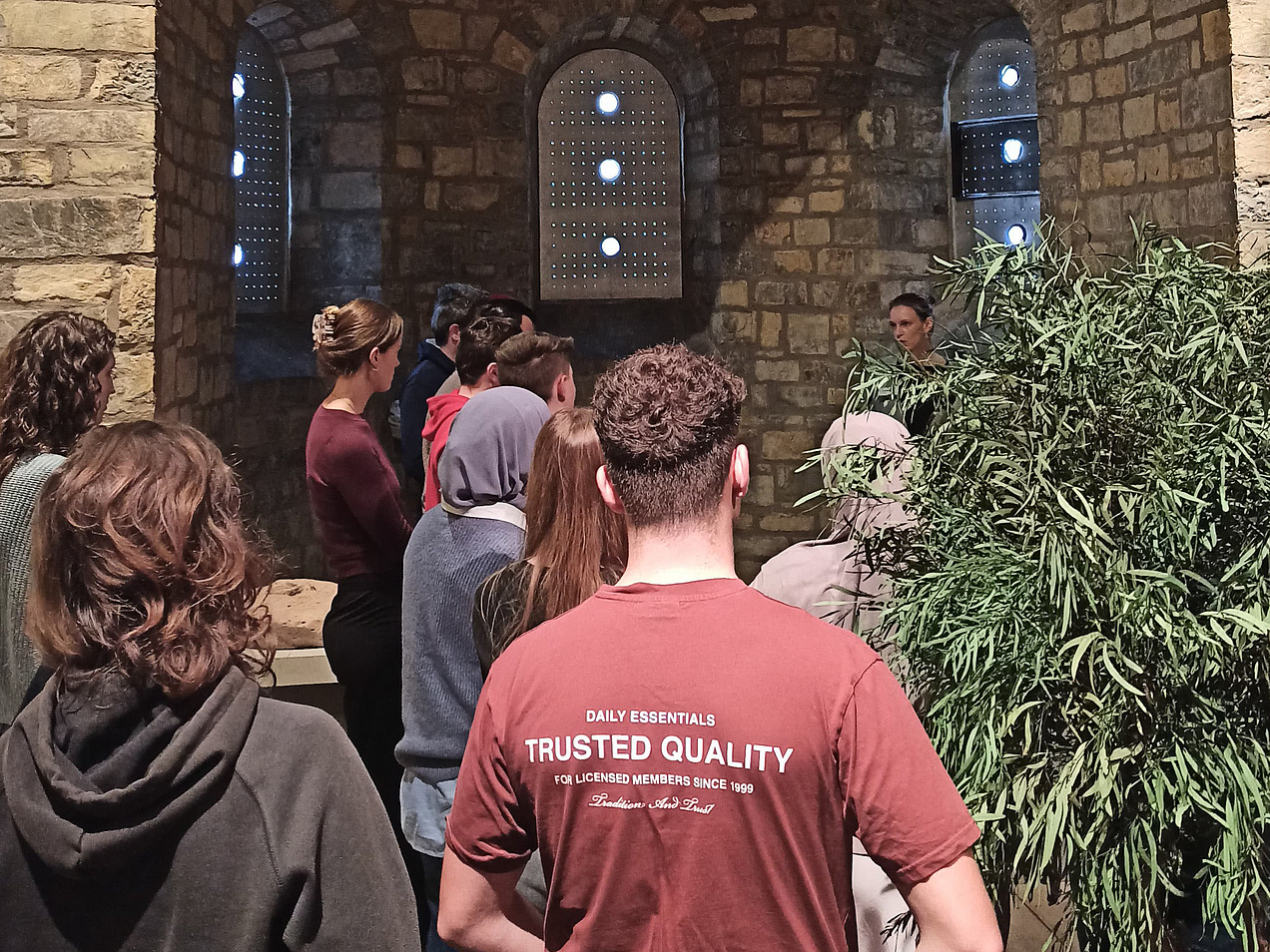 Das Bild zeigt eine Gruppe in einem alten Raum. Die Wände sind aus rauem Stein, rechts ist eine grüne Pflanze.