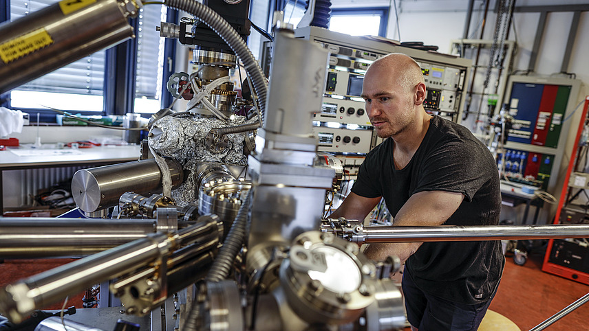 A student sits behind a large assembled device, partially covered with aluminum foil, in a laboratory and observes the process in the device.
