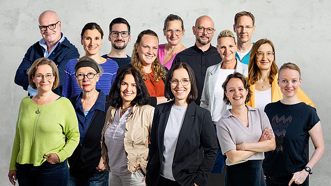 Eine Gruppe Personen steht in einer Gruppe zusammen, alle tragen legere unterschiedliche Kleidung und blicken freundlich in die Kamera.