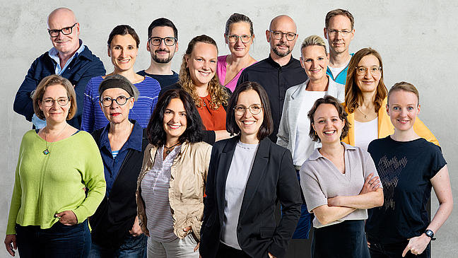 A group of people stand together, all wearing casual, different clothes and looking friendly at the camera.