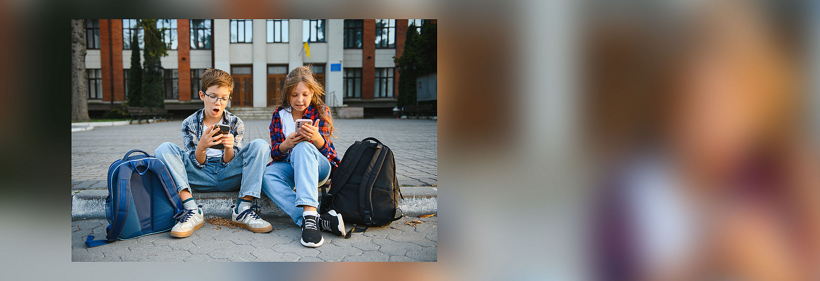 Zwei Kinder im Grundschulalter sitzen auf vor einem schul&auml;hnlichen Geb&auml;ude auf einer Stufe, Sie schauen gespannt auf ihre Smartphones, neben ihnen stehen zwei Rucks&auml;cke.