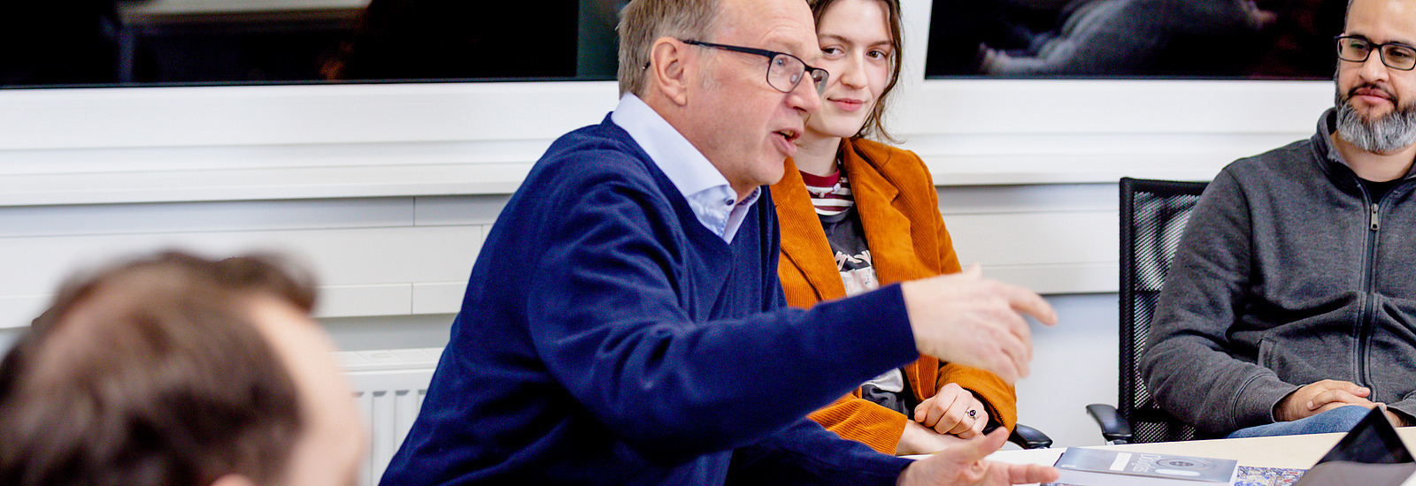 Achim Stephan, emeritierter Philosophieprofessor, spricht gestikulierend mit einer Gruppe von Studierenden. Eine junge Frau in einer orangefarbenen Jacke sitzt neben ihm und hört aufmerksam zu. Weitere Personen sitzen um den Tisch, darunter ein Mann mit Brille und grauem Hoodie. Auf dem Tisch liegen Laptops, Bücher und Notizen. Im Hintergrund spiegelt sich die Szene in einer Fensterscheibe. 
