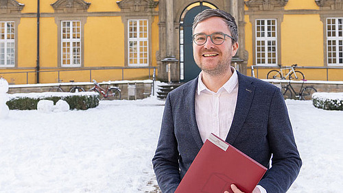 Prof Seitz steht mit einer Mappe in der Hand im Innenhof des Osnabrücker Schlosses. Er lacht in die Kamera.