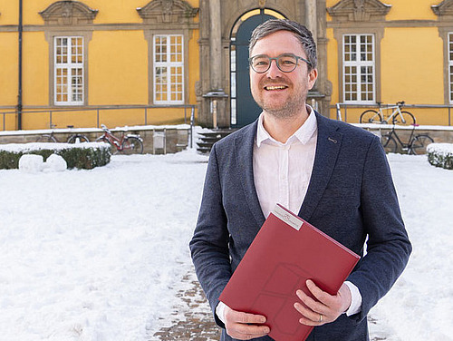 Prof Seitz steht mit einer Mappe in der Hand im Innenhof des Osnabrücker Schlosses. Er lacht in die Kamera.