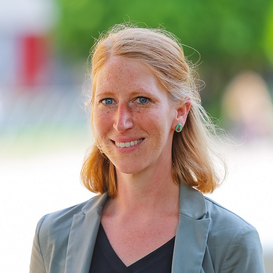 Eine lächelnde Frau mit mittellangem, rötlich-blondem Haar steht im Freien vor einem unscharfen Hintergrund mit grünem Laub. Sie trägt ein schwarzes Oberteil und ein graues Sakko sowie auffällige, grüne Ohrstecker. Ihr Gesicht ist sommersprossig, und sie schaut freundlich in die Kamera.