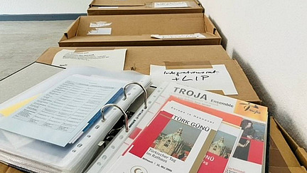 Mehrere geöffnete Pappkartons stehen auf dem Boden, beschriftet mit Zetteln und Notizen. Im Vordergrund liegt ein geöffneter Ordner mit Dokumenten und Prospekten, darunter ein Papier mit der Aufschrift "TROJA Ensemble". Die Kartons sind ordentlich gestapelt, und der Raum hat einen grauen Boden und weiße Wände.
