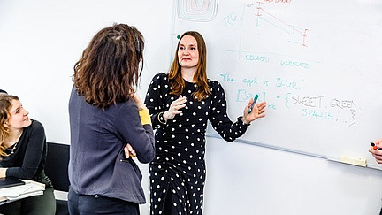 A woman in a black-and-white polka-dotted dress stands in front of a whiteboard, explaining something while holding a green marker. The whiteboard contains diagrams, text, and handwritten notes such as "The apple is sour" and "SWEET GREEN SPINACH...". Another woman in a dark jacket stands opposite her, listening attentively. In the background, other people are sitting at a table, observing the scene.