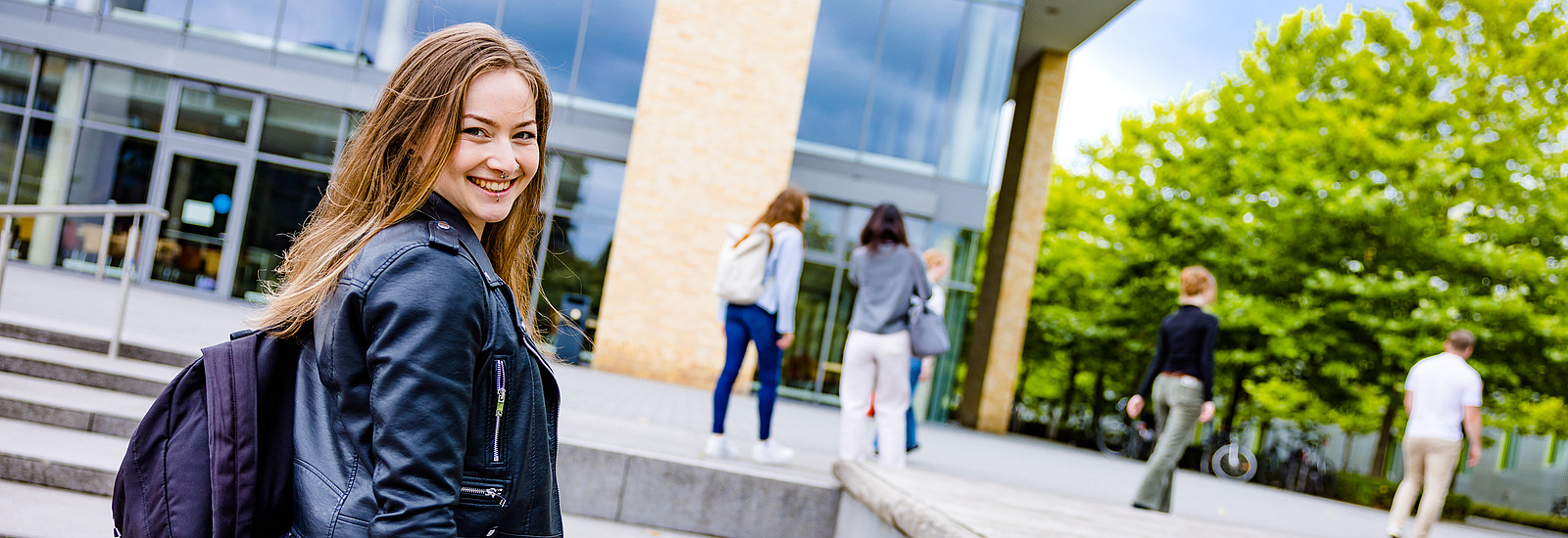 Eine junge Frau auf dem Campus schaut &uuml;ber ihre Schulter lachend in die Kamera, unter dem Arm h&auml;lt sie ein Skateboard. Im Hintergrund sieht man weitere Personen auf ein Geb&auml;ude zugehen.
