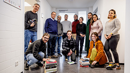 Das Bild ist ein Gruppenfoto der Arbeitsgruppe Philosophie und zeigt zwölf Menschen in einem hell beleuchteten Flur mit weißen Wänden. Sie stehen oder hocken in lockerer Formation zusammen und lächeln in die Kamera. Zwei Personen knien auf dem Boden vor zwei kleinen Stapeln Bücher, während eine stehende Person, Uwe Meyer, im Türrahmen auf der linken Seite ebenfalls Bücher in der Hand hält. Im Hintergrund stehen unter anderem unser Studiendekan Gregor Hörzer, emeritierter Professor Achim Stephan, ehemalige Dozentin Imke von Maur und Doktorand Louis Loock. Die Kleidung der Personen ist vielfältig, von legeren Jeans und Pullovern bis hin zu formelleren Outfits mit Blazern und Schals. Im Hintergrund sind eine Tür mit Glasfenstern und einige an der Wand angebrachte Schilder oder Pläne zu sehen. Die Stimmung ist freundlich und gemeinschaftlich. 