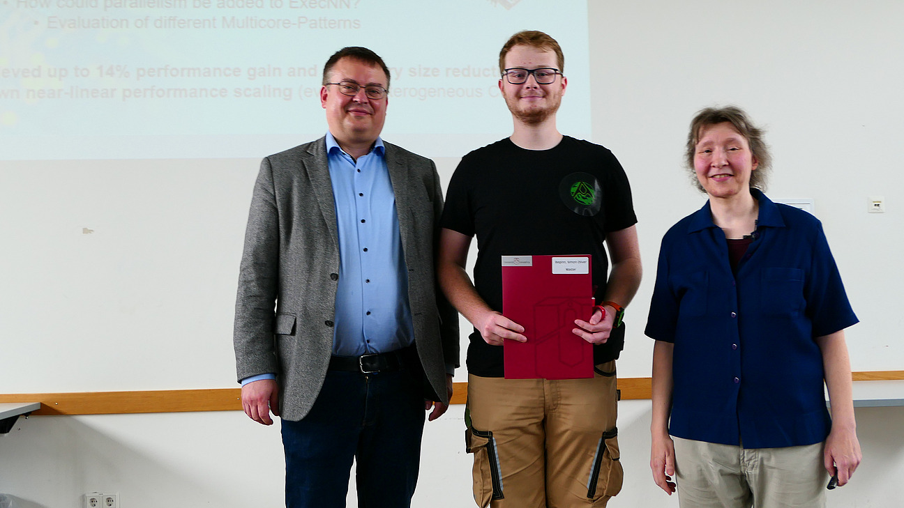Ein Student mit Studiendekanin und Dekan stehen nebeneinander. Der Student hat eine Mappe in der Hand