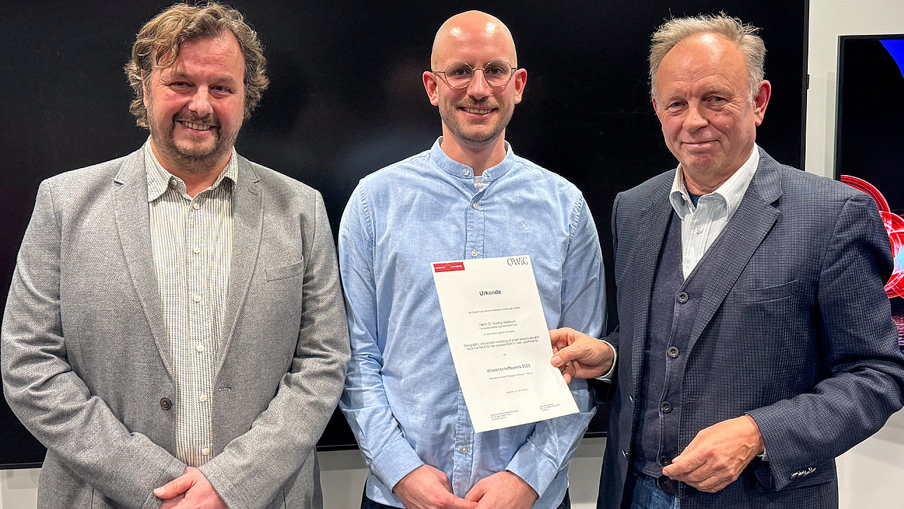 Drei M&auml;nner l&auml;cheln in die Kamera, der rechte Mann h&auml;lt eine Urkunde in der Hand