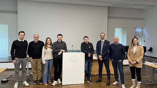 Acht Personen stehen nebeneinander in einem Raum hinter einem Rednerpult. Das Pult steht in der Mitte und tr&auml;gt ein Logo.  Die Personen sind in einer Reihe angeordnet und schauen in Richtung Kamera. Hinter ihnen befindet sich eine gro&szlig;e, helle Wand oder Projektionsfl&auml;che.  Der Raum hat einen Holzboden, und seitlich sind Fenster mit Vorh&auml;ngen zu sehen.