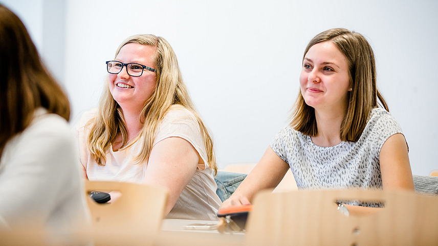 Zwei lächelnde Studentinnen sitzen in einem Seminarraum und schauen nach Vorne.