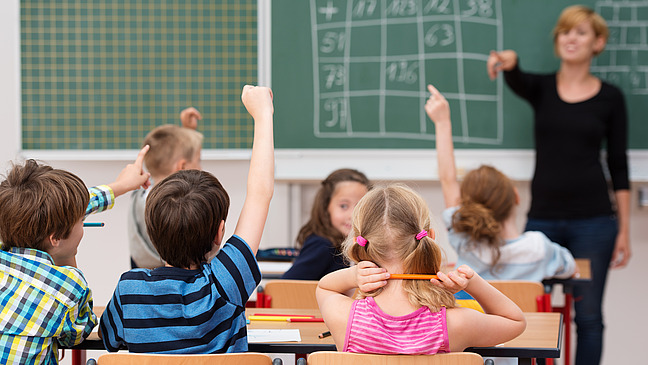 In a classroom, several children are sitting at their desks with their hands raised to ask a question. A boy in a checked shirt and a girl in a pink T-shirt are clearly visible. In the foreground, the teacher is standing at the blackboard, talking and looking in the direction of the pupils.