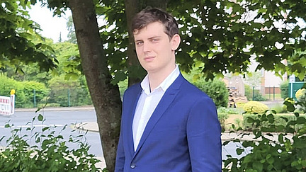 Ein junger Mann in einem blauen Anzug und weißem Hemd steht draußen vor einem Baum. Er hat kurze, braune Haare und einen ernsthaften Gesichtsausdruck. Die Umgebung ist grün mit Blättern und Pflanzen im Hintergrund, eine Straße ist ebenfalls sichtbar. 