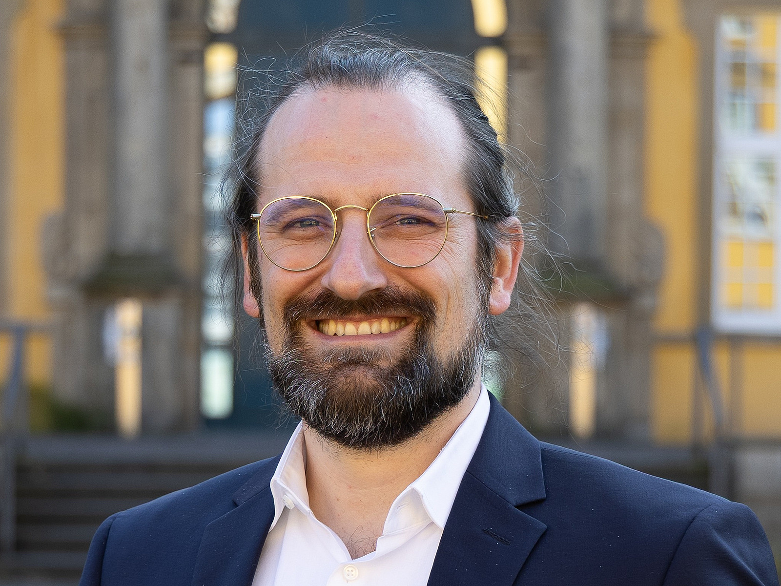 Foto von einem Herren mit Brille, Bart und langen, zusammengebunden Harren in einem Anzug. Er lächelt.