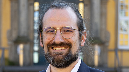 Foto von einem Herren mit Brille, Bart und langen, zusammengebunden Harren in einem Anzug. Er lächelt.