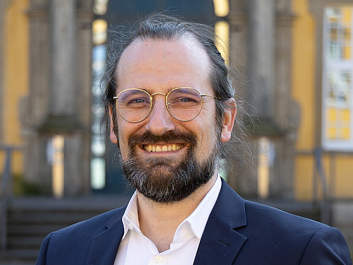 Foto von einem Herren mit Brille, Bart und langen, zusammengebunden Harren in einem Anzug. Er lächelt.