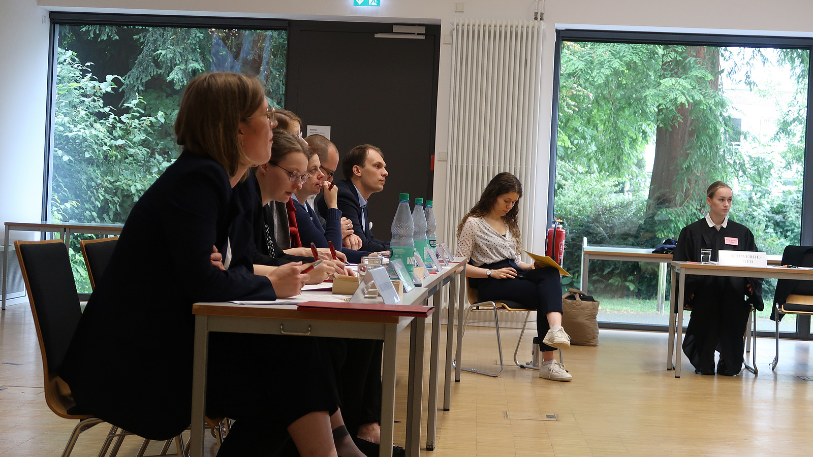 Tischreihe mit aufmerksam zuh&ouml;renden Personen, daneben eine Schriftf&uuml;hrerin und ein junge Frau in schwarzer Robe