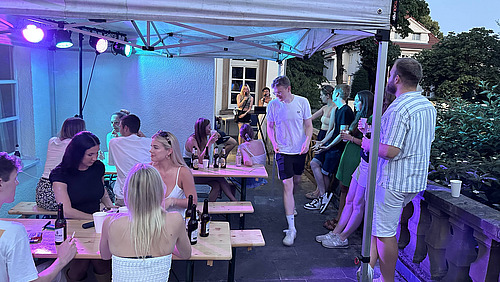 Mehrere Personen sitzen und stehen an Holztischen unter einem &uuml;berdachten Bereich. Einige sitzen auf B&auml;nken, andere lehnen oder stehen daneben. Auf den Tischen stehen Flaschen und Gl&auml;ser.  Ein Mann in heller Kleidung geht durch den Raum zwischen den Tischen. &Uuml;ber den Personen sind Lichtquellen angebracht, die den Bereich in verschiedene Farben tauchen.  Im Hintergrund ist eine Wand sowie ein Durchgang zu einem weiteren Bereich zu sehen, in dem sich ebenfalls Personen aufhalten.