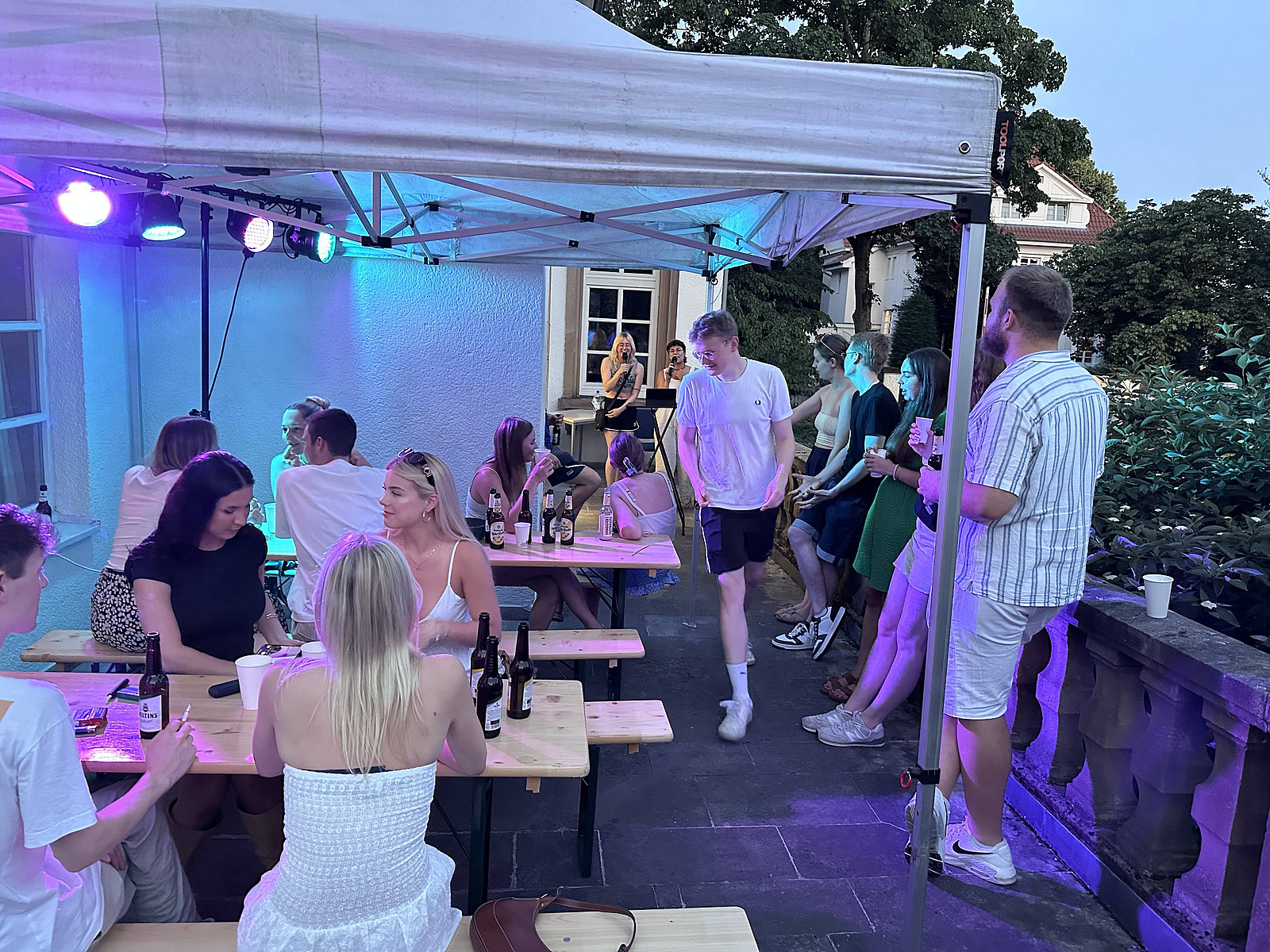 Mehrere Personen sitzen und stehen an Holztischen unter einem &uuml;berdachten Bereich. Einige sitzen auf B&auml;nken, andere lehnen oder stehen daneben. Auf den Tischen stehen Flaschen und Gl&auml;ser.  Ein Mann in heller Kleidung geht durch den Raum zwischen den Tischen. &Uuml;ber den Personen sind Lichtquellen angebracht, die den Bereich in verschiedene Farben tauchen.  Im Hintergrund ist eine Wand sowie ein Durchgang zu einem weiteren Bereich zu sehen, in dem sich ebenfalls Personen aufhalten.