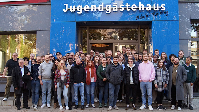 Eine große Personengruppe steht vor einem Gebäude. Über dem Gebäudeeingang ist der Schriftzug "Jugendgästehaus Adolph Kolping" zu lesen.
